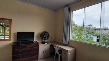 Quarto com televisão sobre cômoda de madeira, um ventilador e janela com vista para área verde e edifícios.