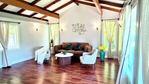 A spacious living room with a wooden floor, a brown sofa, a white armchair, and a round coffee table. There is a large window with curtains and a wall-mounted light fixture.