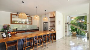 A modern kitchen with a wooden island, bar stools, pendant lights, and a view of a staircase leading to a balcony.