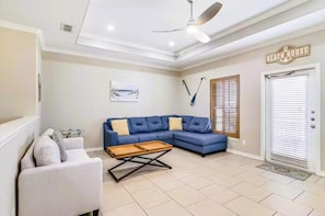 A living room with a blue sofa, a white armchair, a wooden coffee table, a ceiling fan, and a door labeled "BEACH HOUSE".