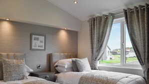 Ein Hotelzimmer mit zwei Betten, einem fenster mit Vorhängen, einem Kopfteil mit einer Leuchte und einem gerahmten Bild an der Wand.