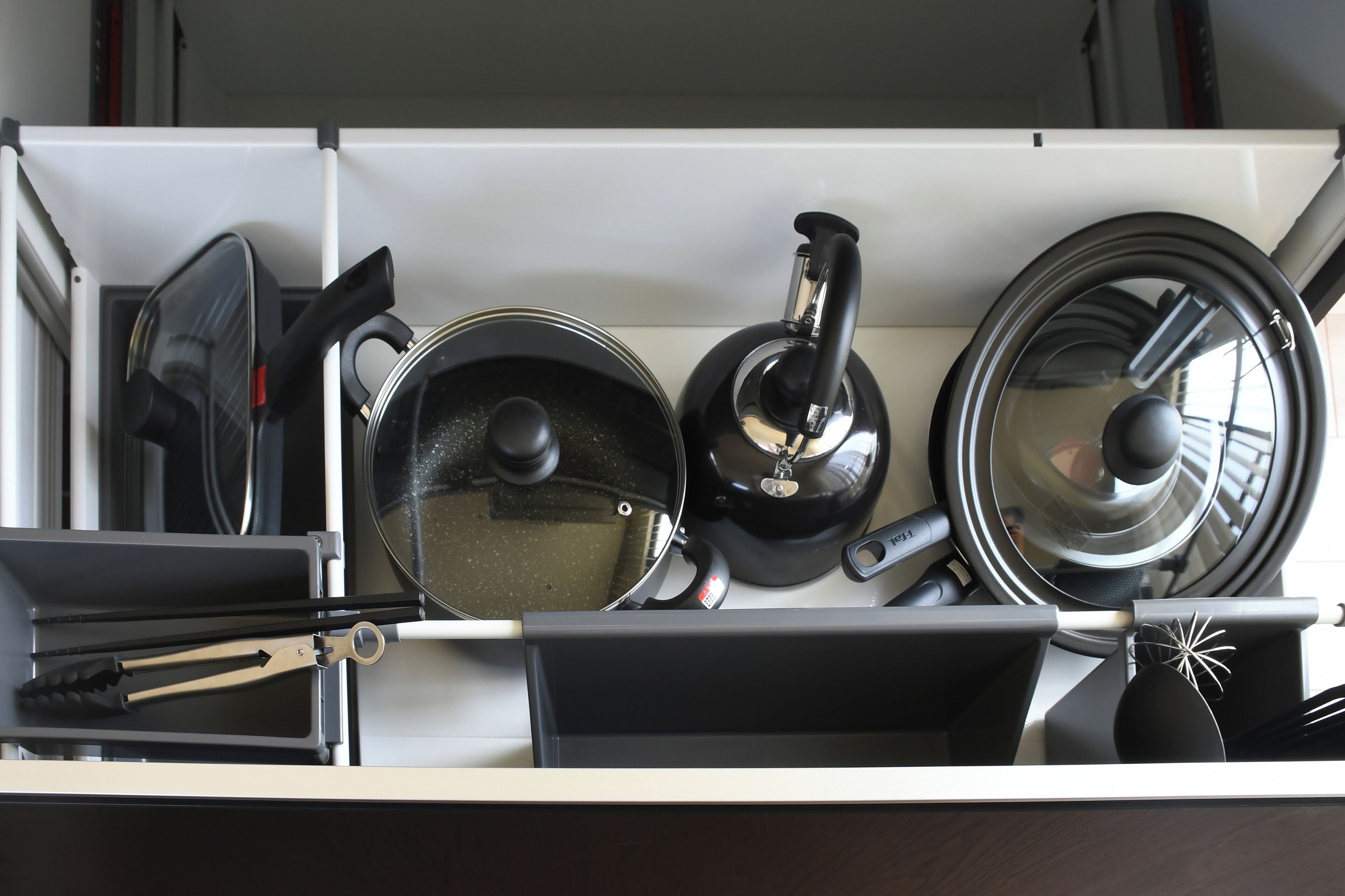 A kitchen drawer with various utensils and cookware, including a kettle, pots, and tongs.