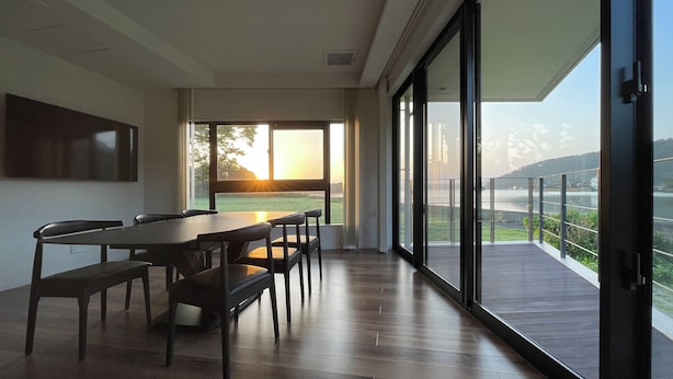 Un espace de repas moderne, doté d’une grande table et de chaises, de larges fenêtres offrant une vue sur un plan d’eau et des collines, ainsi que d’un balcon avec une balustrade.