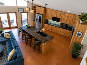 A modern kitchen with wooden cabinets, a central island with bar stools, and a dining area with a blue sofa and chairs.