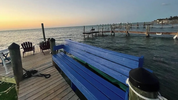 Un muelle de madera con un banco y sillas azules, que se adentra en aguas tranquilas bajo un cielo despejado.