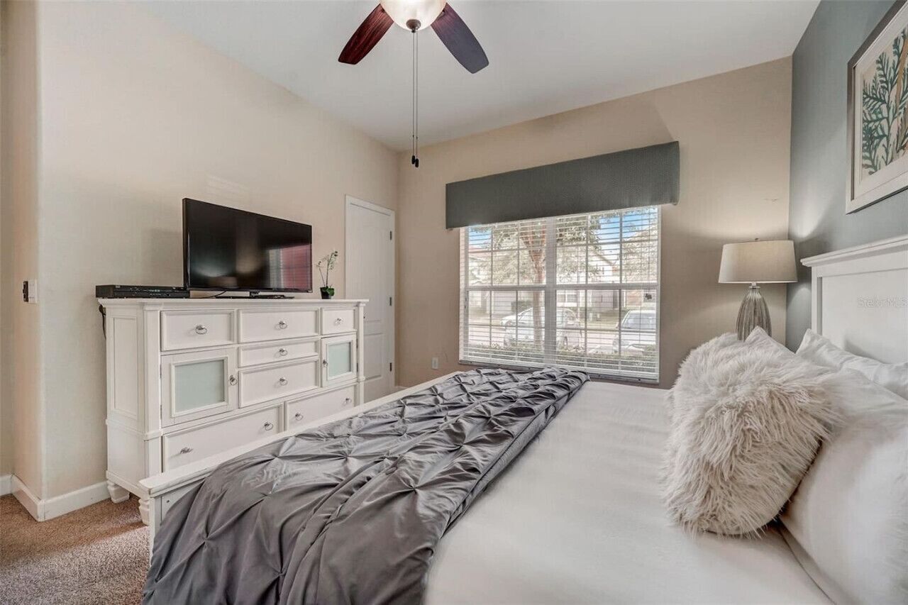 A bedroom with a bed, a television on a dresser, a window with blinds, and a ceiling fan.