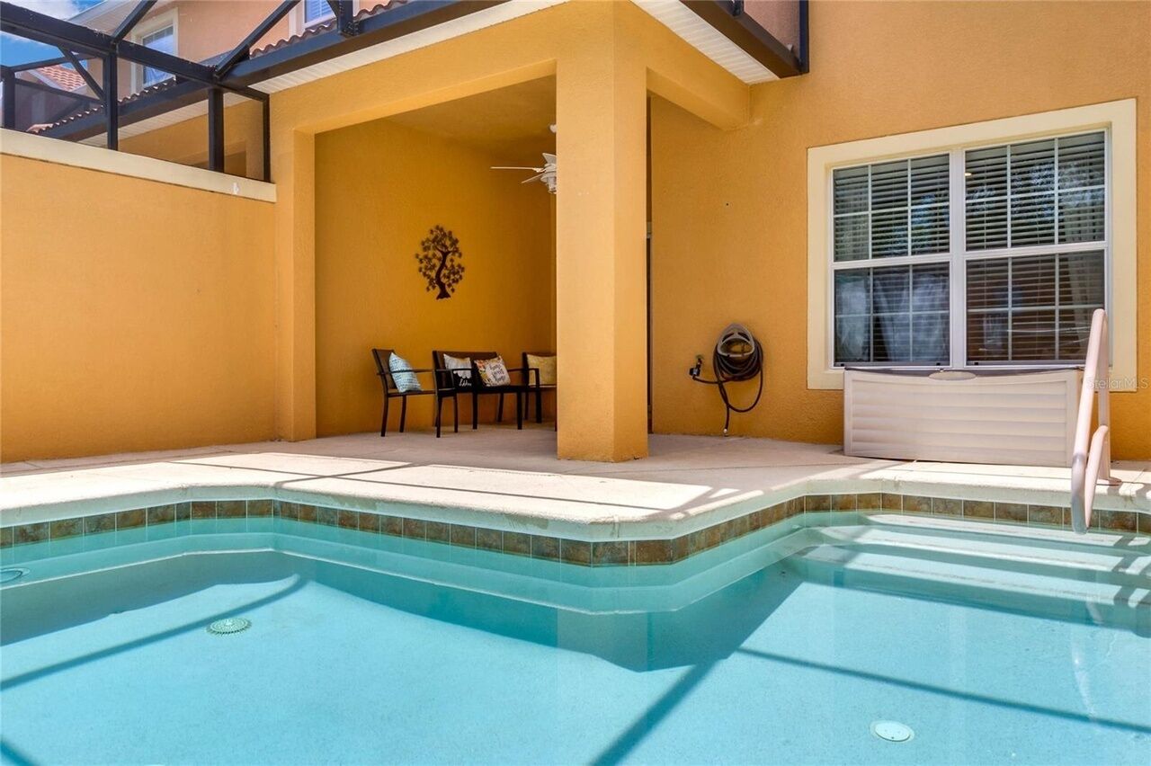 A swimming pool with a tiled edge, a covered seating area with chairs, and a window with blinds.