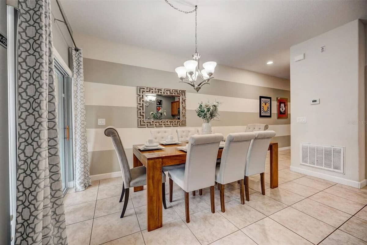 A dining area with a wooden table, white upholstered chairs, a chandelier, a mirror, and a decorative wall piece.