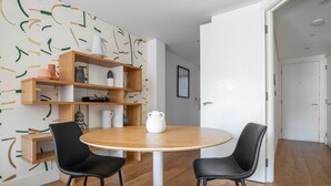 A dining area with a round wooden table, two black chairs, and a white pitcher. There is a wooden shelf unit with various items on it, and a white door in the background. - Bright Aldgate 1BR, close to Train & Liverpool Street, by Blueground (London)