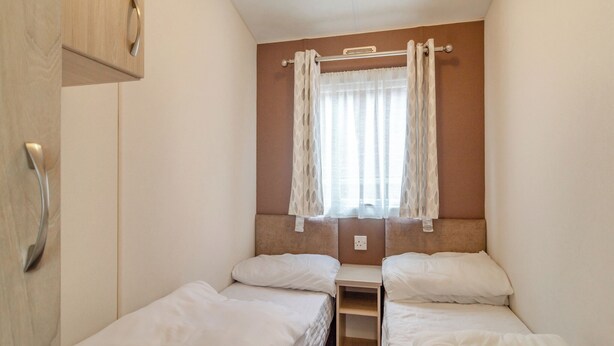 A small, single-room hotel room with two beds, a wooden wardrobe, a window with sheer curtains, and a wall-mounted shelf. - Castaways Oyster 10 (Pet) (Bacton-on-Sea)