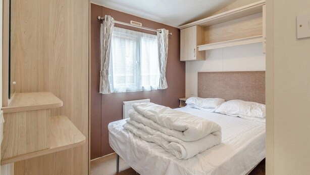 A bedroom with a bed, a window with curtains, a radiator, and built-in wooden shelves. - Castaways Oyster 10 (Pet) (Bacton-on-Sea)