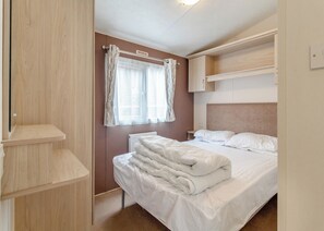 A bedroom with a bed, a window with curtains, a radiator, and built-in wooden shelves. - Castaways Oyster 10 (Pet) (Bacton-on-Sea)