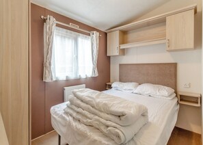 A bedroom with a bed, curtains, a headboard, a bedside table, and a built-in wardrobe. - Castaways Oyster 10 (Pet) (Bacton-on-Sea)