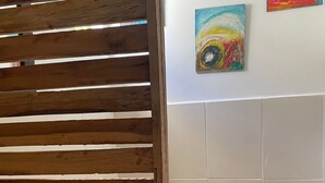 A bathroom with a wooden slatted partition, a toilet, a trash bin, and two abstract paintings on the wall.