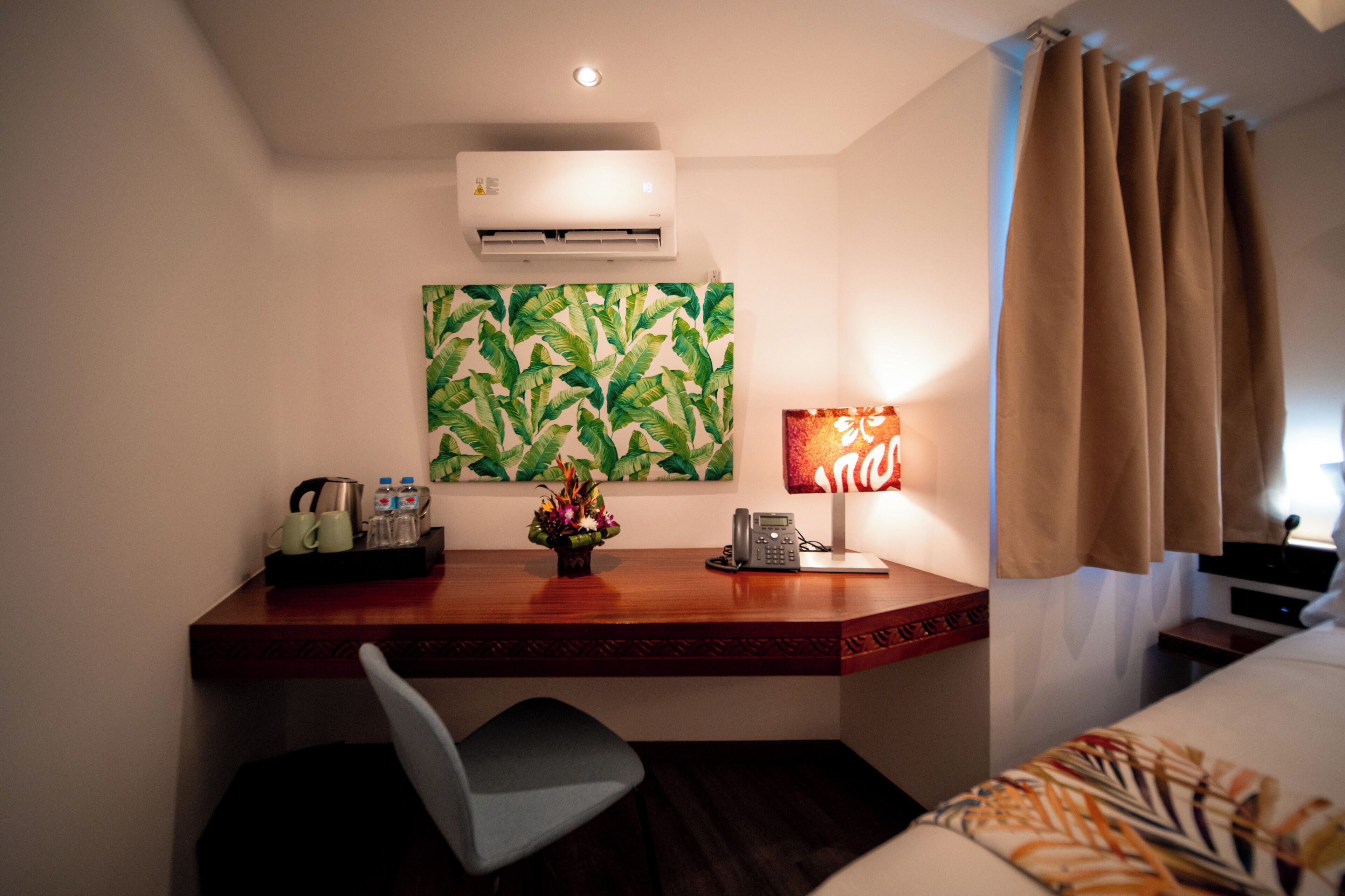 Une chambre d’hôtel équipée d’un bureau, d’une chaise, d’un climatiseur, d’œuvres d’art, d’une lampe et de rideaux.