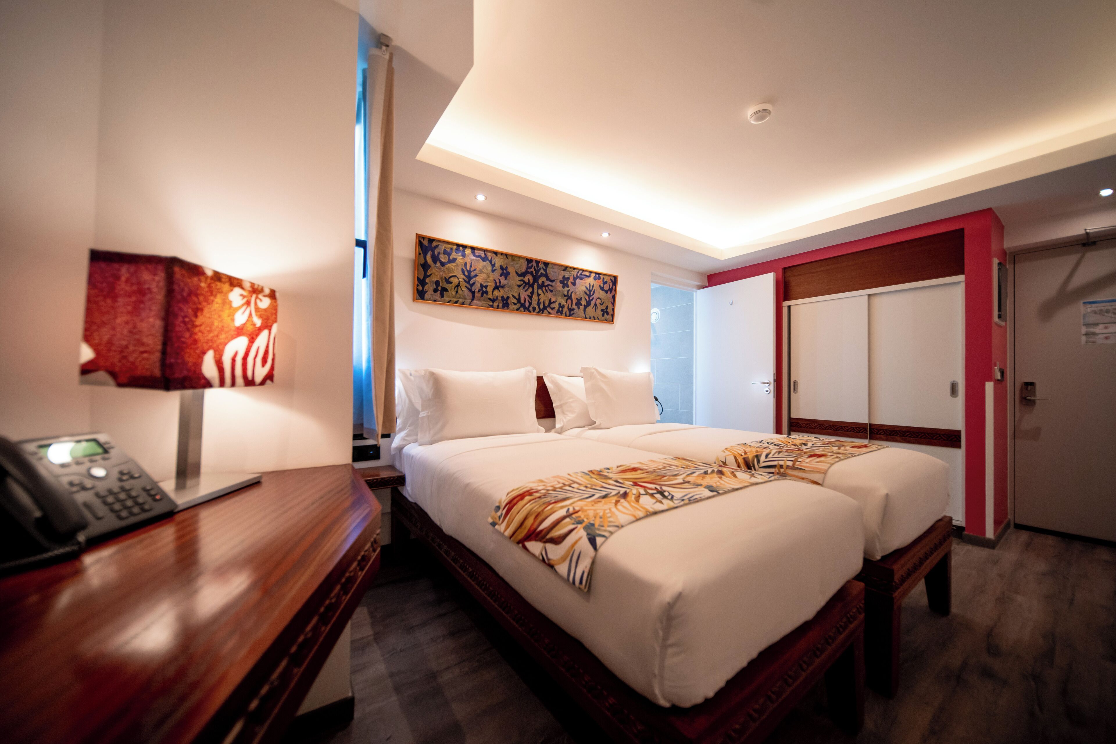 Habitación de hotel con una cama grande, un escritorio con teléfono, un cuadro en la pared y una puerta que da a otra habitación.