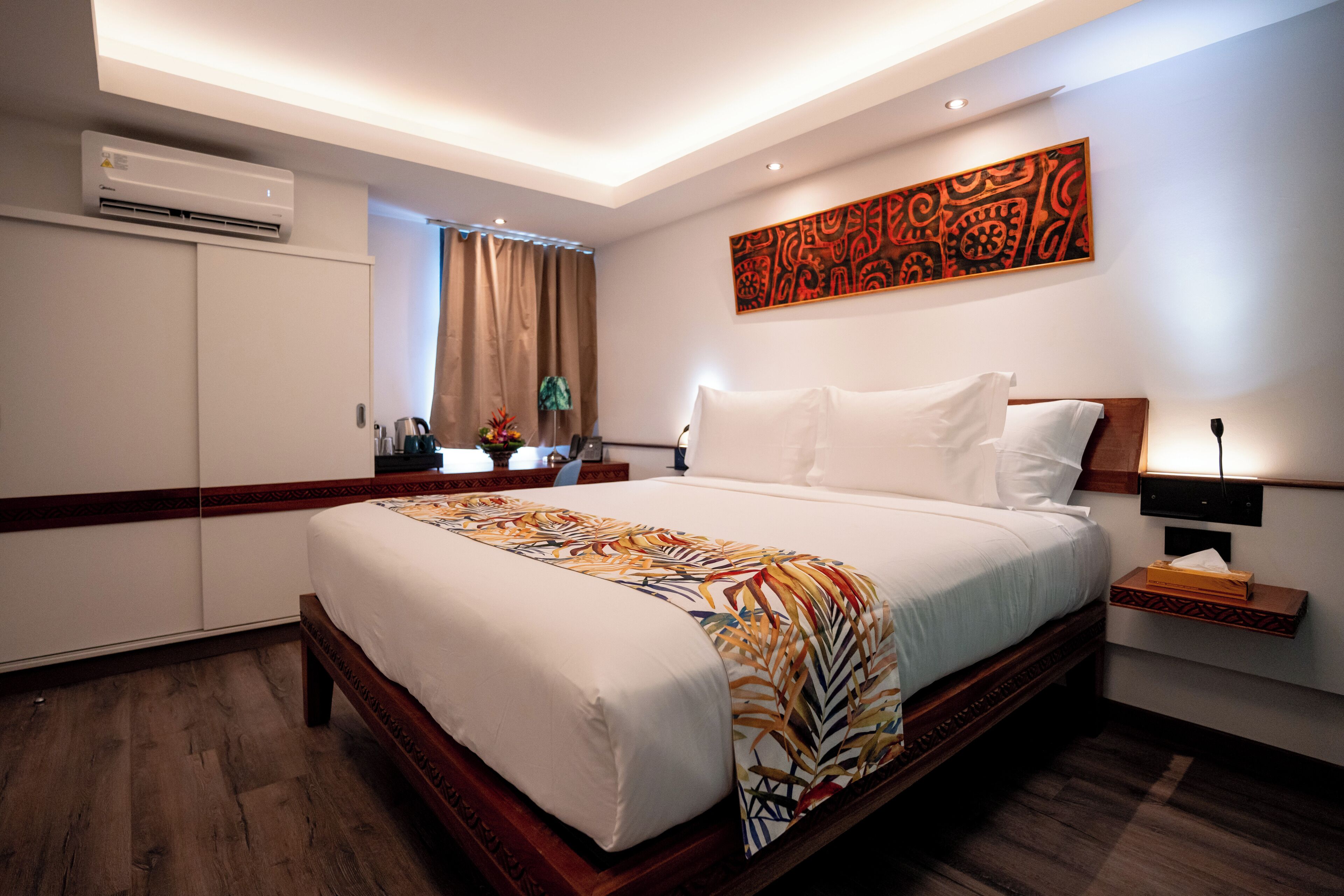 A hotel room with a large bed, a nightstand, a wall-mounted air conditioner, and a decorative wall piece.