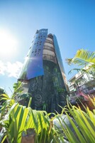 Un edificio alto y cilíndrico con una fachada única, perforada, y una base decorada con vegetación.