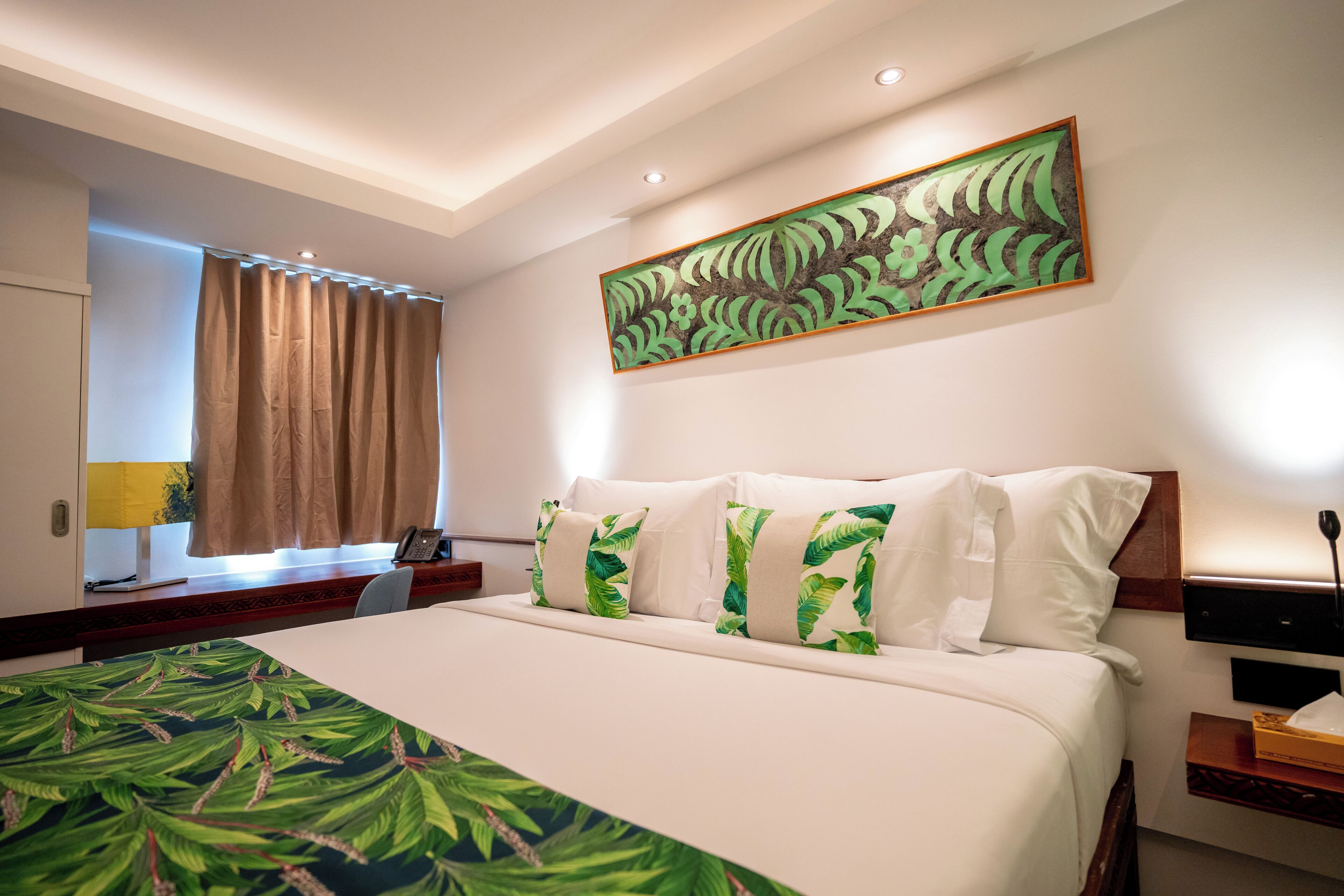 A hotel room with a large bed, a desk with a lamp, a painting on the wall, and a window with curtains.