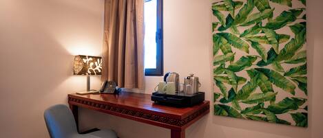 A hotel room with a wooden desk, a chair, a window with curtains, a wall-mounted air conditioner, and a large leaf-patterned artwork.