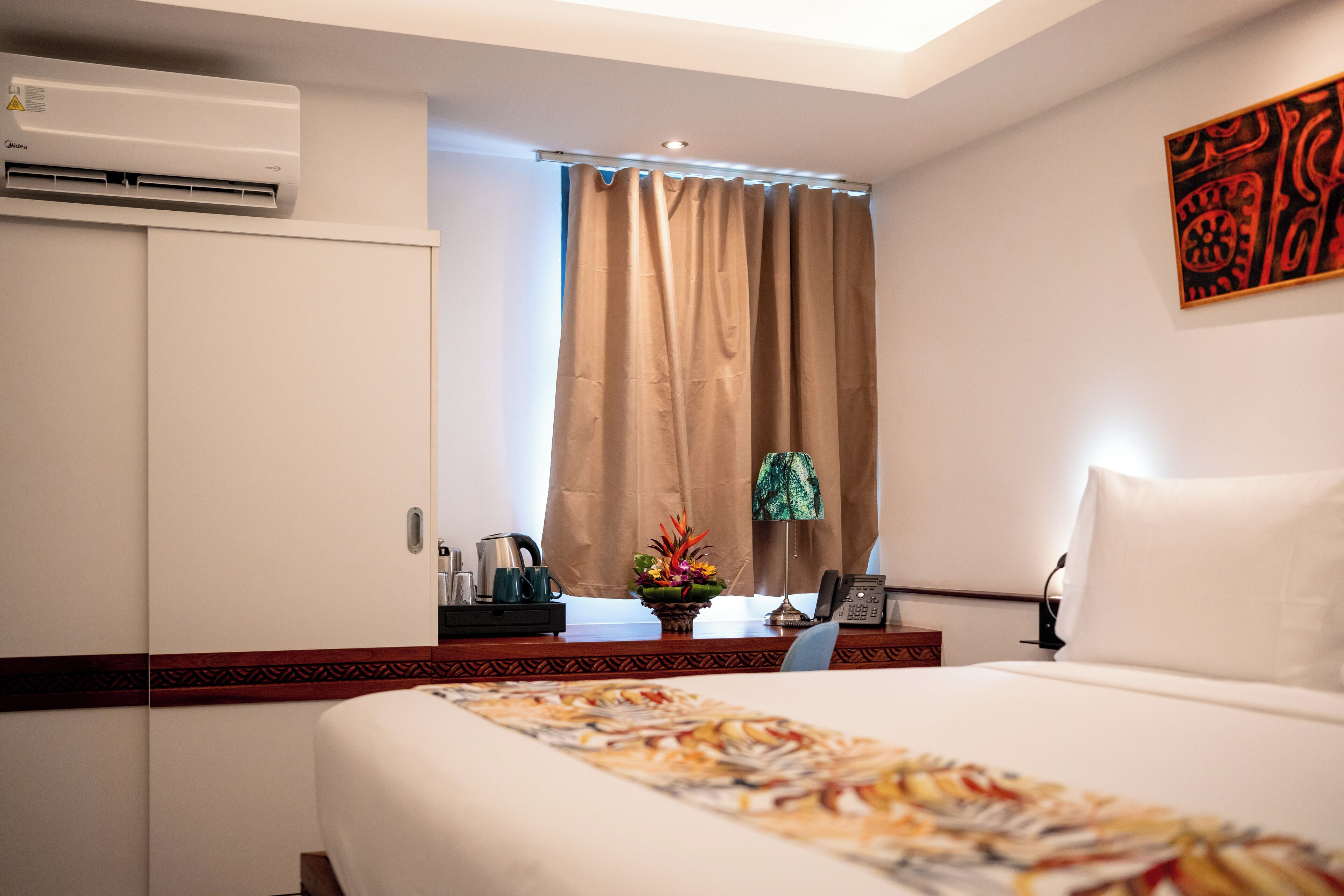 Una habitación de hotel con cama, escritorio con teléfono, cafetera, un jarrón con flores y un equipo de aire acondicionado.