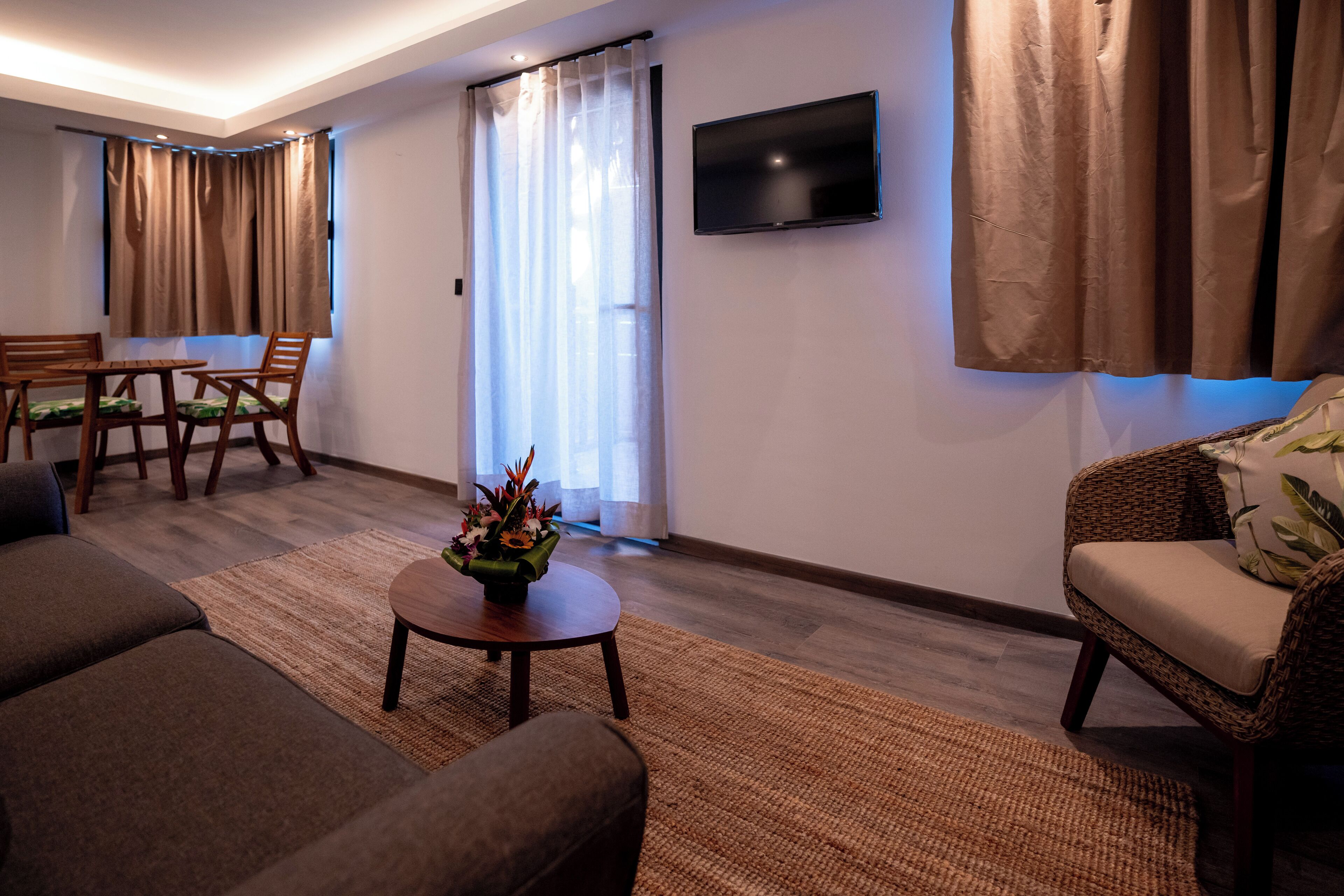 A living room with a sofa, a coffee table with a flower arrangement, a dining area with a table and chairs, and a TV mounted on the wall.