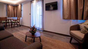 A living room with a sofa, a coffee table with a flower arrangement, a dining area with a table and chairs, and a TV mounted on the wall.