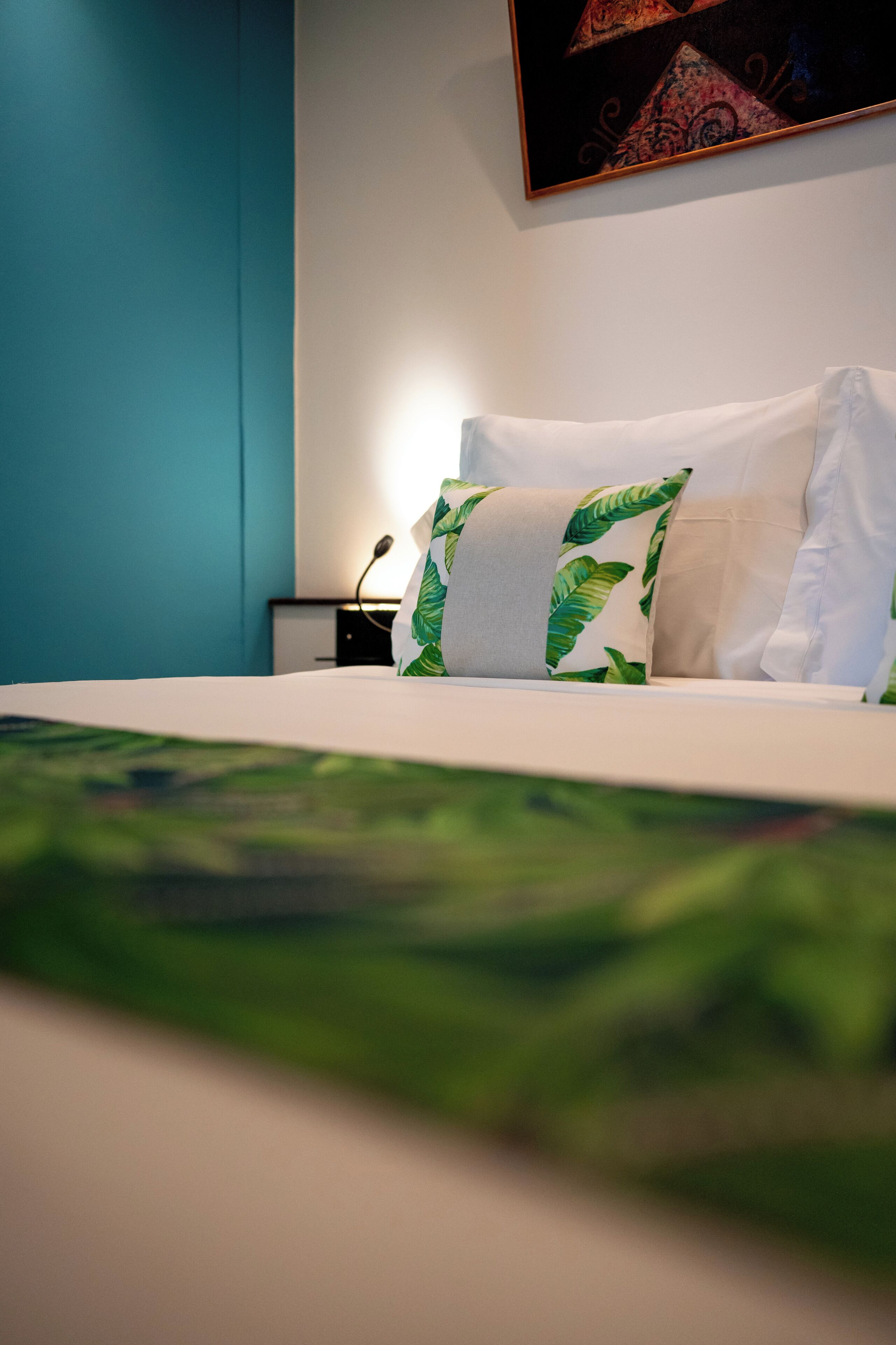 Un lit bien fait, recouvert d’une couvre-lit à motifs de feuilles vertes, avec des oreillers blancs et un tableau accroché au mur au-dessus du lit.