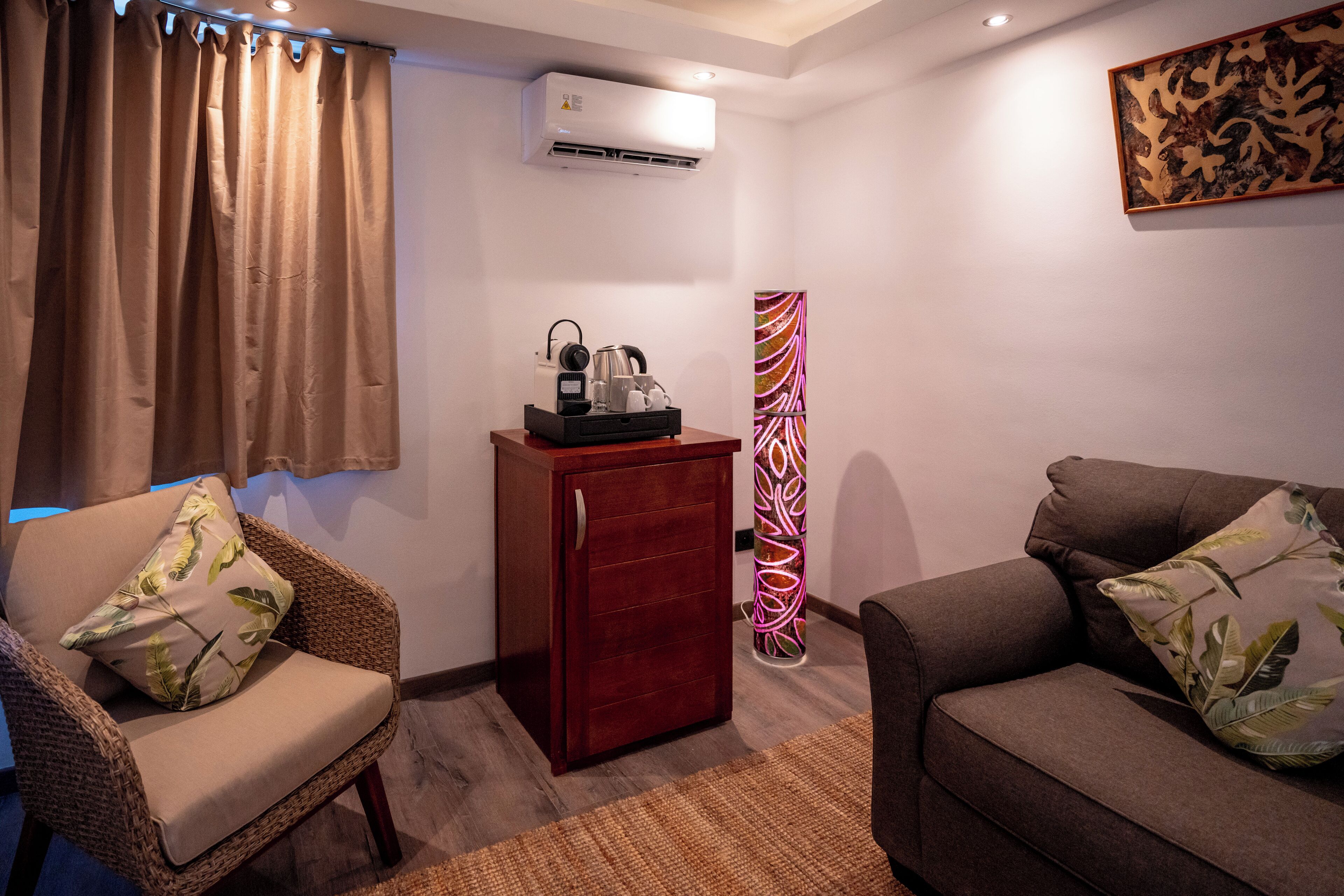 A living room with a brown sofa, a wicker chair, a small wooden cabinet, and a wall-mounted air conditioner.