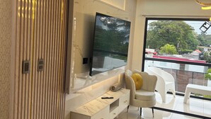 A modern living room with a large flat-screen TV mounted on a marble wall, a white sofa, and a glass door leading to a balcony with a view of greenery and buildings.