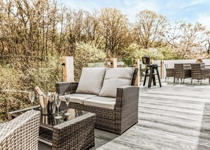 A wicker sofa with cushions, a table with chairs, and a bottle of champagne on a deck.