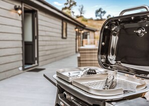A portable grill with utensils and a tray, placed on a patio with a house in the background.
