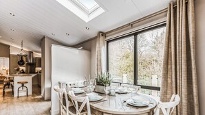 A dining area with a round wooden table set for four, surrounded by white chairs, a large window with curtains, and a skylight above.