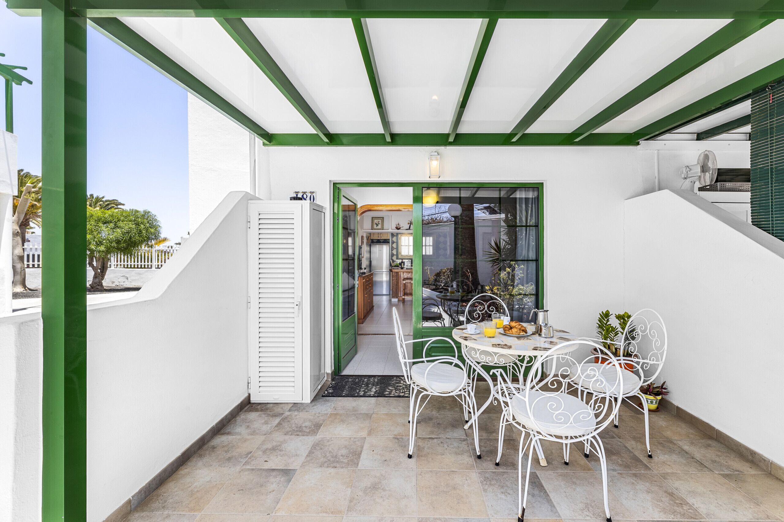 Une terrasse couverte avec une table et des chaises, une porte donnant sur un espace intérieur et un plafond bordé de verdure.