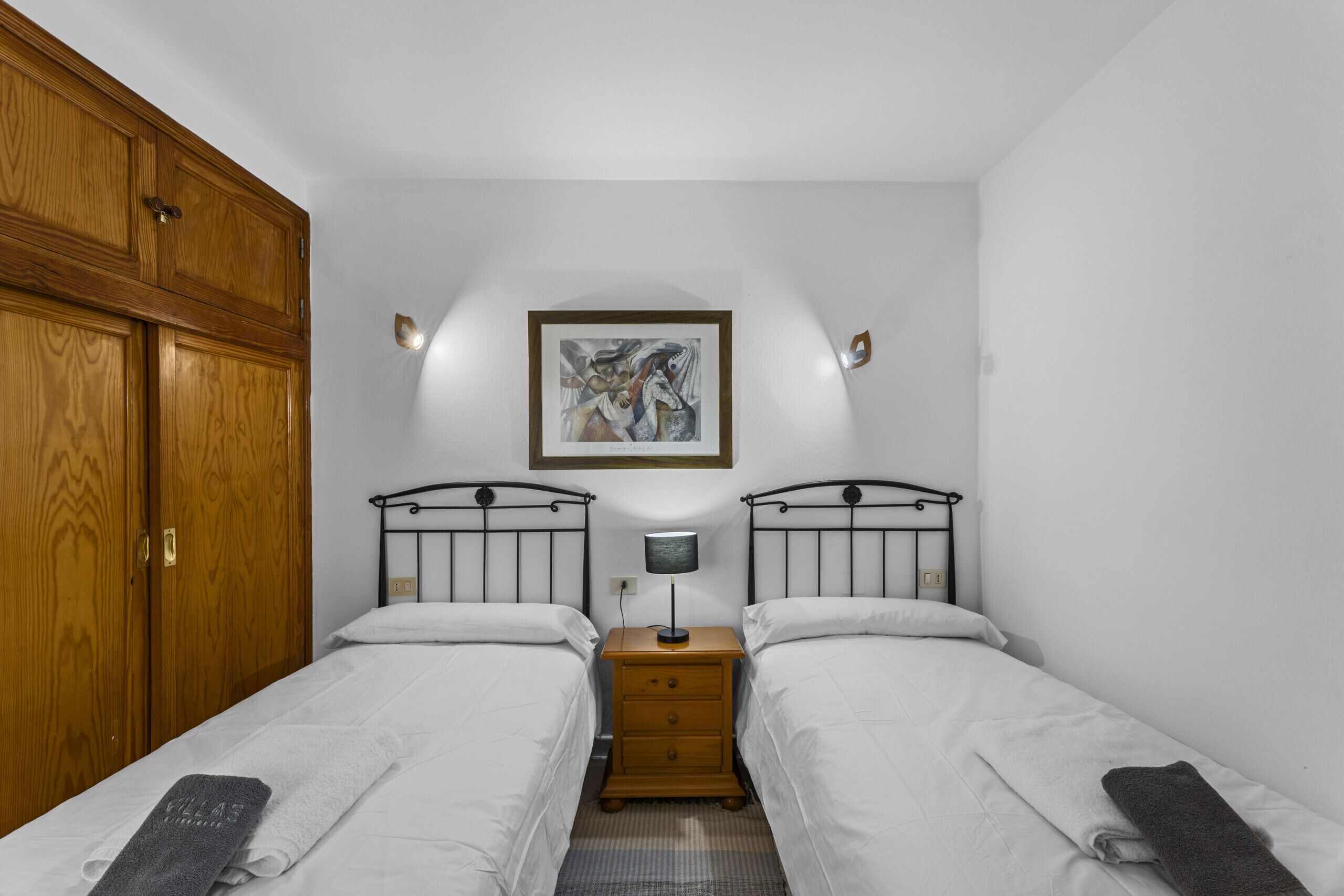 A bedroom with two beds, a wooden wardrobe, a nightstand with a lamp, and a framed picture on the wall.