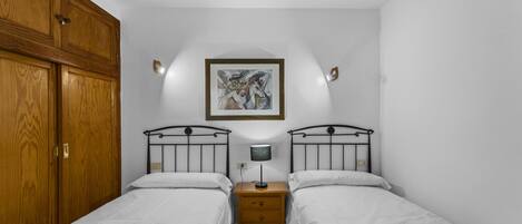 A bedroom with two beds, a wooden wardrobe, a nightstand with a lamp, and a framed picture on the wall.