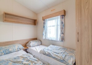 A bedroom with two beds, a window with curtains, a wooden shelf, and a wardrobe. - Castaways Oyster 6 (Pet) (Bacton-on-Sea)