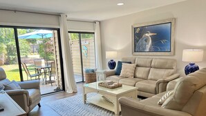 Un salon spacieux avec un canapé beige, une table basse en verre et une vue sur l’extérieur grâce à de grandes portes-fenêtres coulissantes.