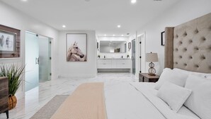 Un dormitorio moderno con una cama grande, una mesita de noche con lámpara, un espejo y un baño a simple vista al fondo.