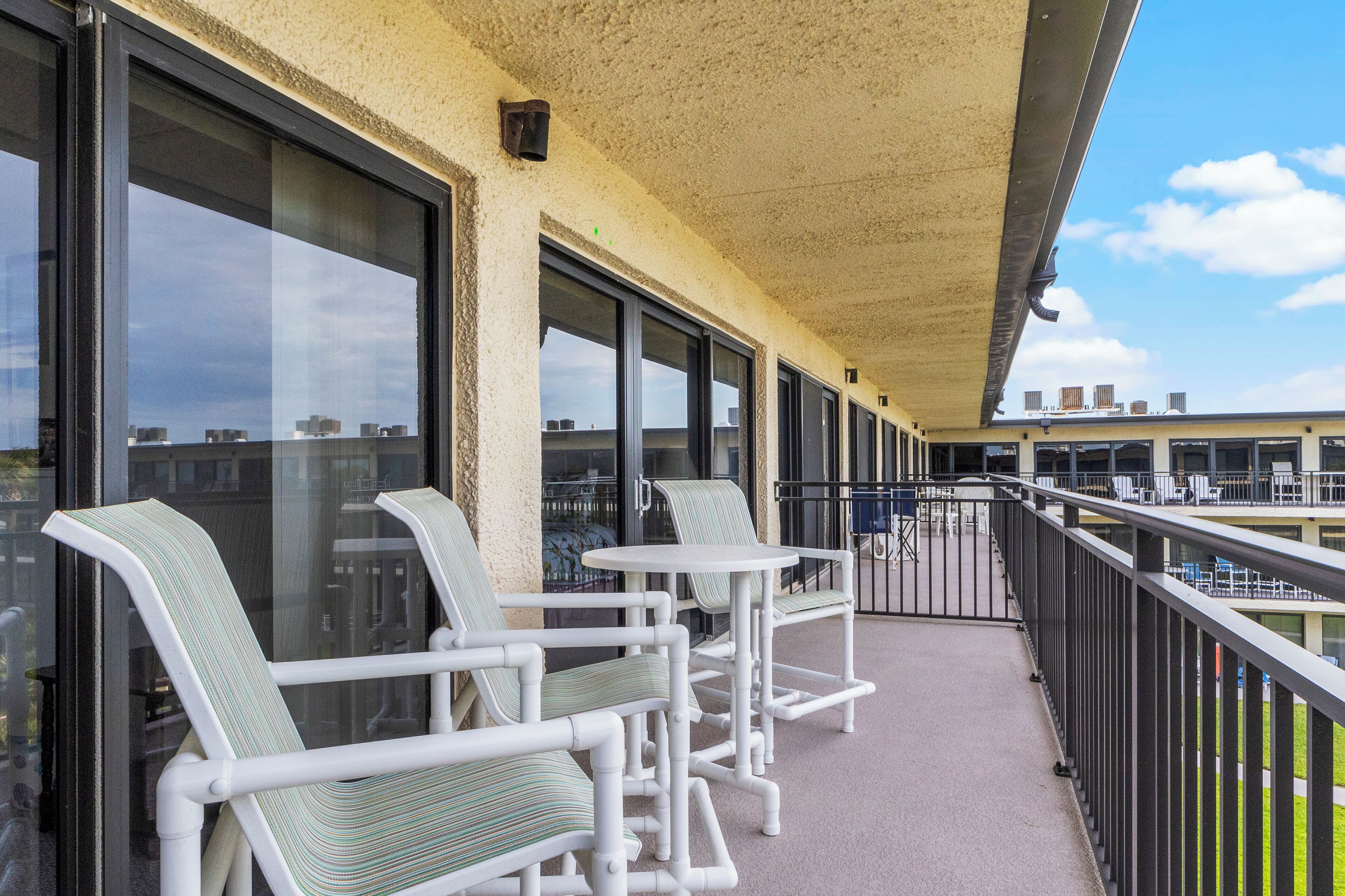 A balcony with white chairs and tables, large windows, and a view of buildings in the background.