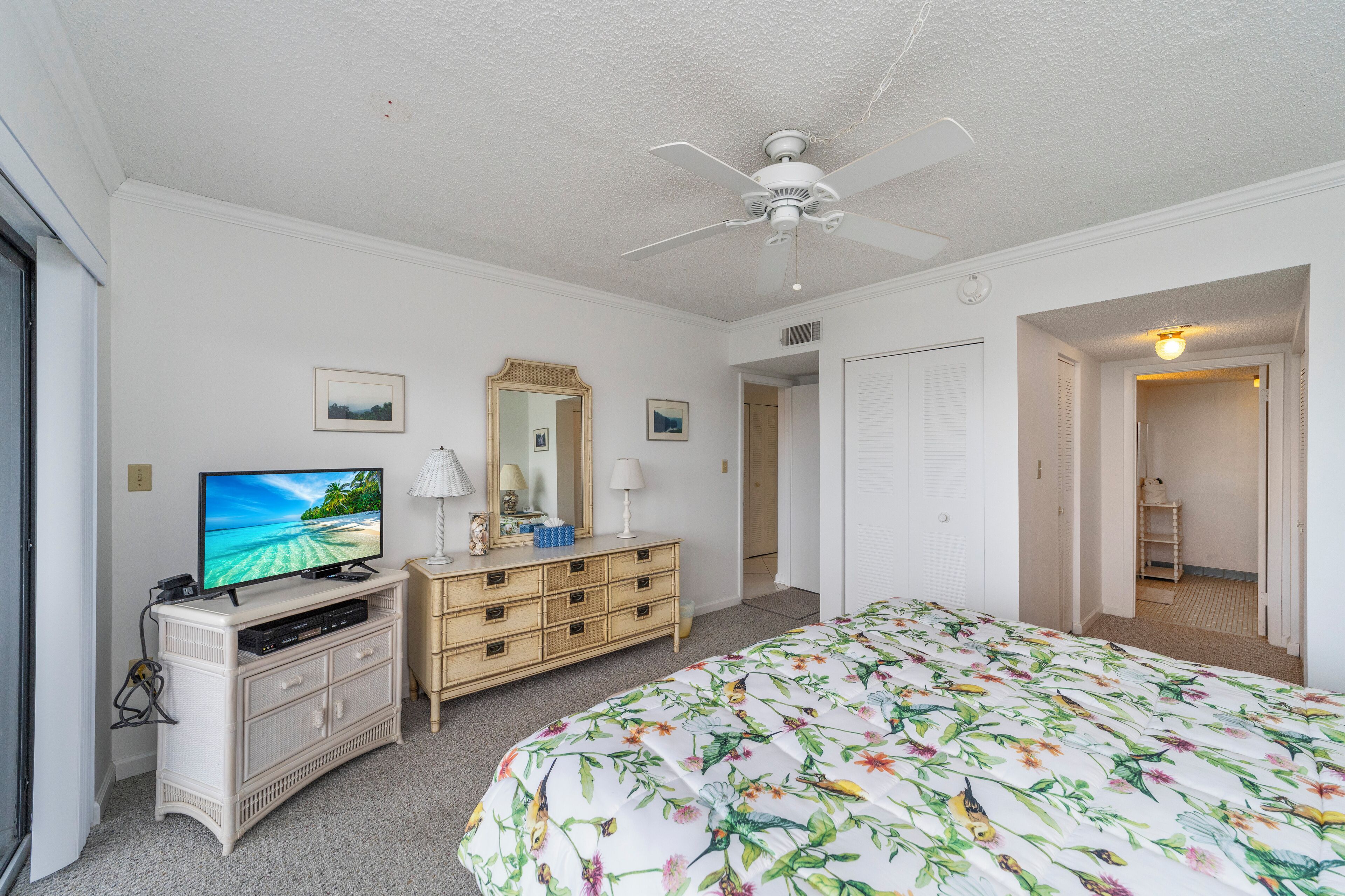 Un dormitorio con una cama, un televisor sobre un aparador, un ventilador de techo y una puerta que da a otra habitación.
