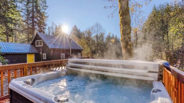 Un bain à remous entouré d’une terrasse en bois, avec une maison et une forêt en arrière-plan.
