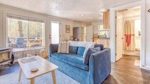 Un salon chaleureux avec un canapé bleu, une petite table en bois et une cheminée. On aperçoit un coin cuisine avec des placards et un réfrigérateur, le tout visible à travers une porte ouverte.