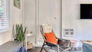 Un salon moderne doté d’une grande fenêtre, d’un canapé, d’un fauteuil avec un coussin moelleux, d’une petite table d’appoint et d’un téléviseur fixé au mur.