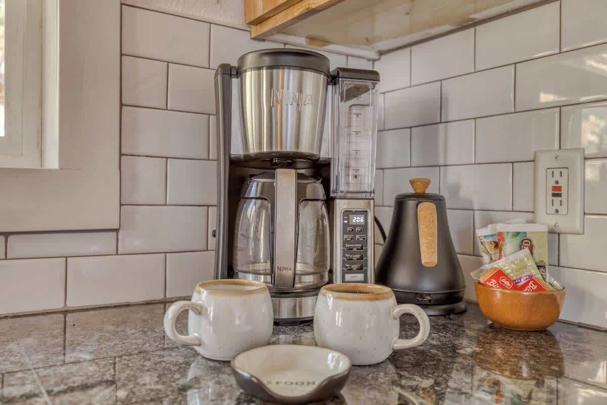 Una cocina con una cafetera, dos tazas, una cuchara y un bol sobre la encimera.