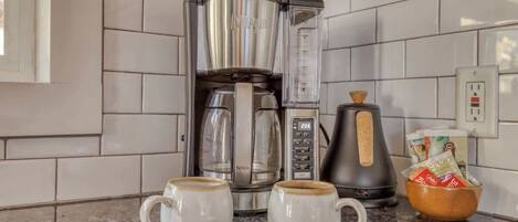 Una cocina con una cafetera, dos tazas, una cuchara y un bol sobre la encimera.