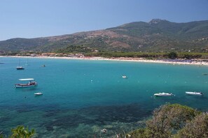 Blick auf die Küste mit Booten, klarem Wasser und einem Strand, im Hintergrund sind Gebäude zu sehen.