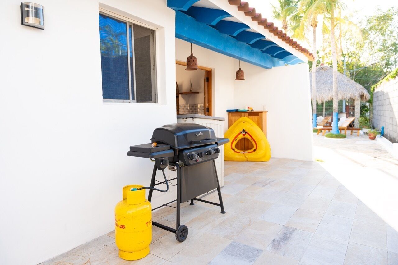 Une terrasse extérieure équipée d’un barbecue, d’une bouteille de gaz jaune et d’une structure à toit de chaume.