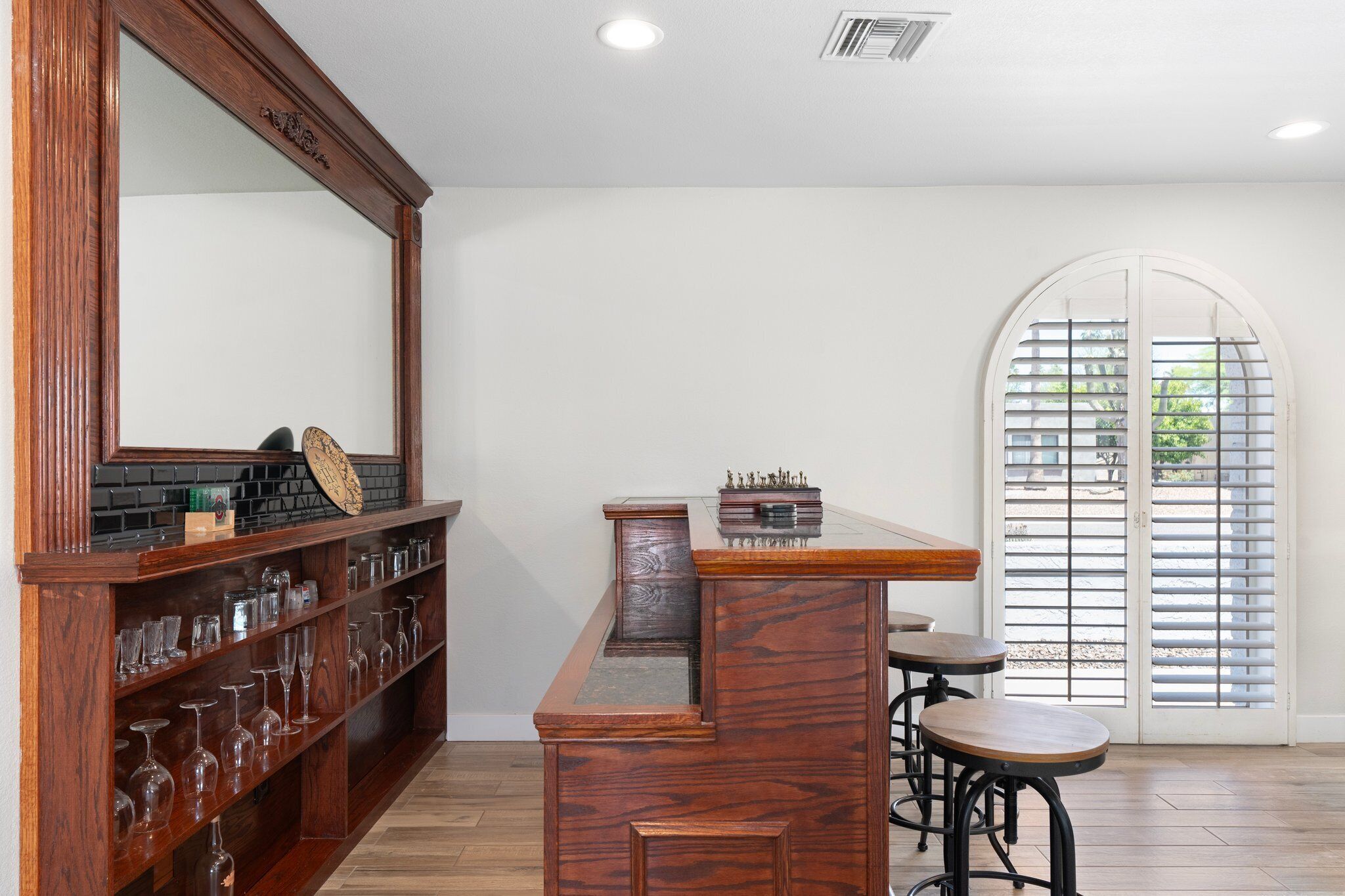 A bar area with a wooden counter, shelves with glasses, and a large mirror. There are two stools and a window with blinds.