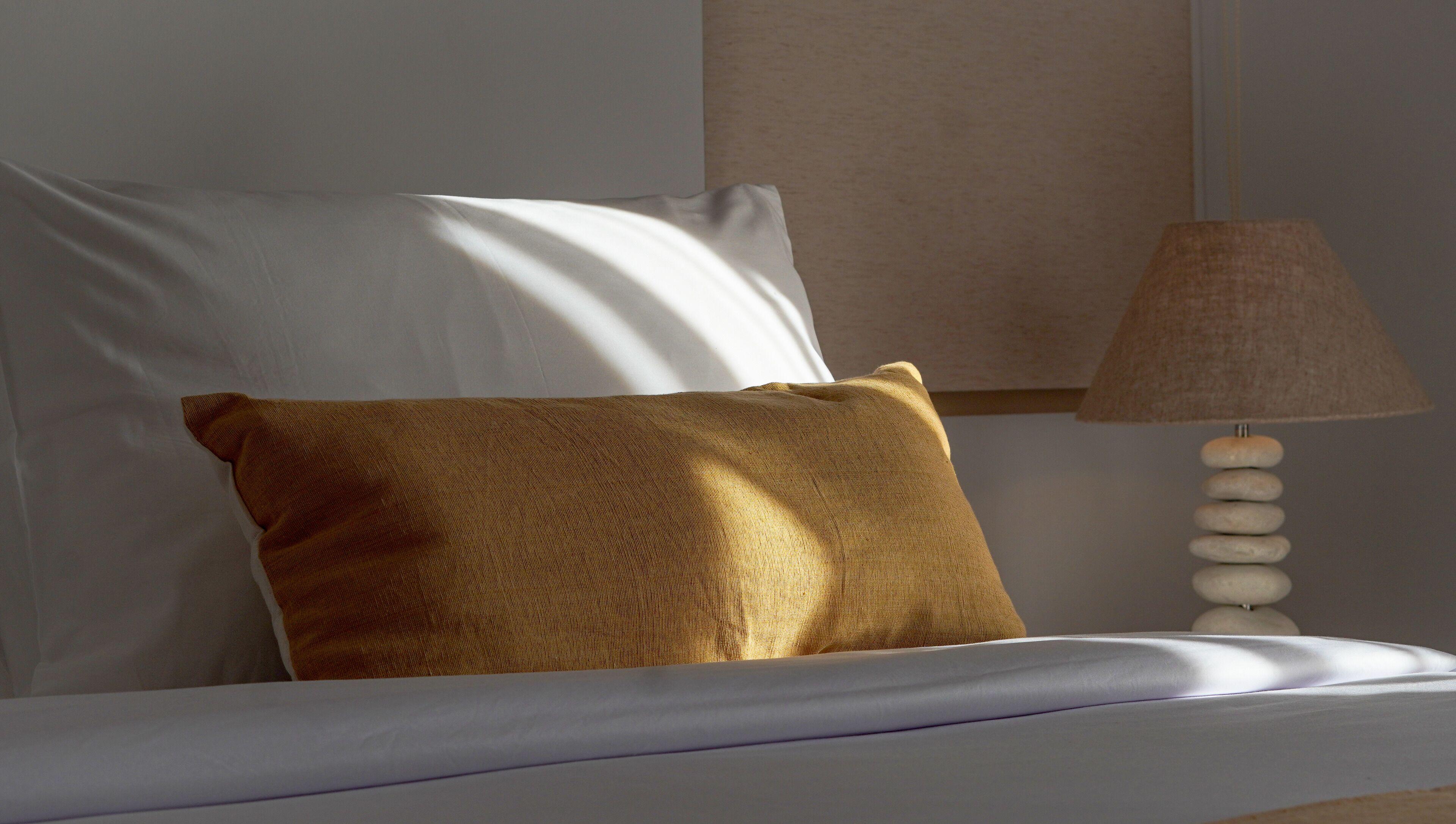 A bed with white sheets and a brown pillow, a beige wall, and a lamp with a beige shade.